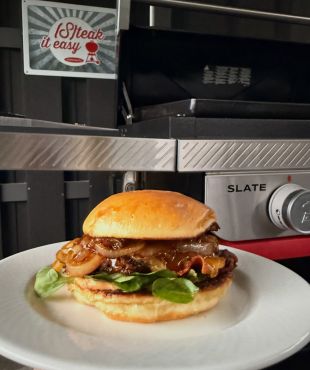 Nahaufnahme eines Burgers auf einem weißen Teller. Der Burger besteht aus einem goldbraunen Brötchen, saftigem Fleisch-Patty, geschmolzenem Käse, karamellisierten Zwiebeln und frischen grünen Blättern. Im Hintergrund steht ein Grill.