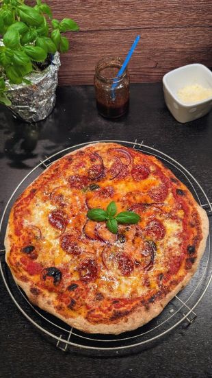 Frisch gebackene Pizza auf einem runden Abkühlgitter. Die Pizza ist belegt mit geschmolzenem Käse, Tomatensauce, Salami, roten Zwiebelringen und einigen schwarzen Oliven. In der Mitte liegt ein frisches Basilikumblatt als Garnitur.