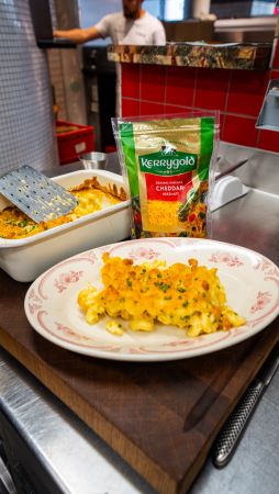 Gericht mit cremigem Mac and Cheese, serviert auf einem weißen Teller mit rotem Muster. Im Hintergrund steht eine Auflaufform mit weiterer Portion, daneben eine Packung Kerrygold Cheddar gerieben.