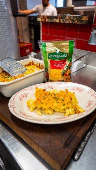 Gericht mit cremigem Mac and Cheese, serviert auf einem weißen Teller mit rotem Muster. Im Hintergrund steht eine Auflaufform mit weiterer Portion, daneben eine Packung Kerrygold Cheddar gerieben.