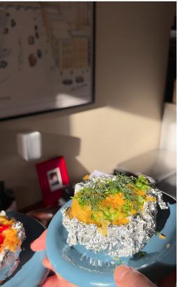 Nahaufnahme einer Ofenkartoffel in Alufolie, serviert auf einem blauen Teller. Die Kartoffel ist mit geschmolzenem Käse, frischen Kräutern und grünen Zwiebelstücken garniert.