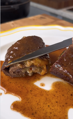 Nahaufnahme eines aufgeschnittenen Rinderroulade-Gerichts auf einem weißen Teller mit gelbem Rand. Die Roulade ist mit einer dunklen, glänzenden Bratensoße überzogen und wird gerade mit einem Messer aufgeschnitten, sodass die Füllung aus Speck, Zwiebeln und geschmolzenem Käse sichtbar wird.