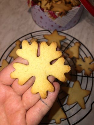 Hand hält ein frisch gebackenes Plätzchen in Schneeflockenform. Im Hintergrund liegt ein rundes Abkühlgitter mit weiteren Weihnachtsplätzchen in Stern- und Tannenbaumform.