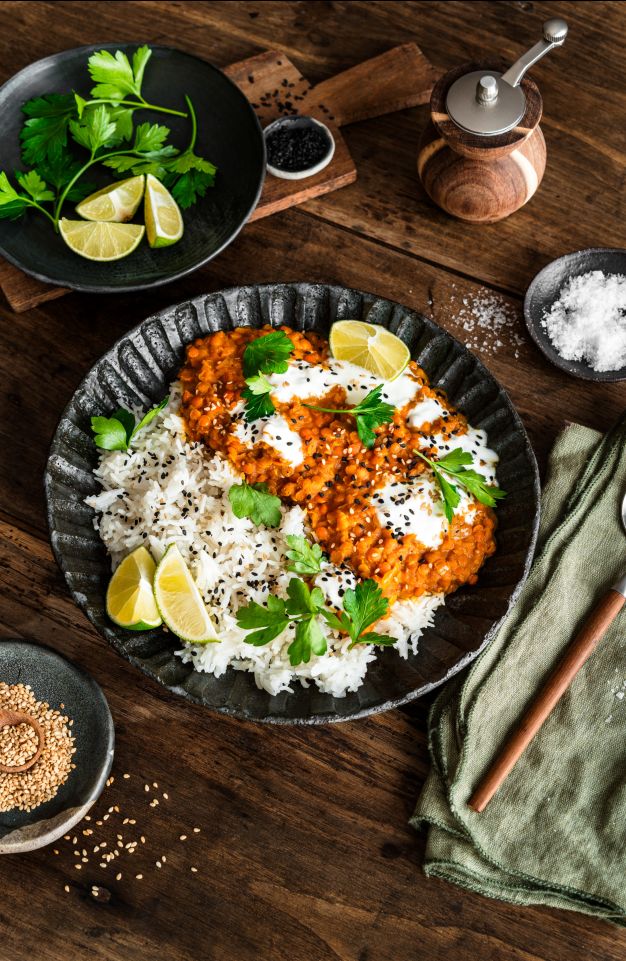 Eine Schüssel mit weißem Reis, garniert mit Linsen-Curry, Joghurt, Sesamkörnern, Limettenspalten und frischen Kräutern, steht auf einem rustikalen Holztisch. Daneben liegen ein Löffel, eine Serviette und Zutaten wie Sesamkörnern, Limette und Gemüse.