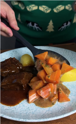 Nahaufnahme eines Tellers mit geschmortem Fleisch in dunkler Sauce, gekochten Kartoffeln und einem Löffel mit glasierten Karotten in Buttersoße.