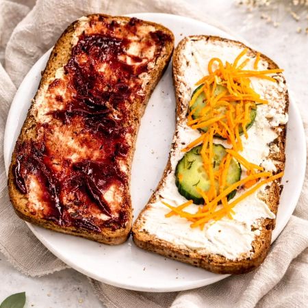 Zwei Scheiben Vollkornbrot auf einem weißen Teller: die linke bestrichen mit Frischkäse und roter Marmelade, die rechte mit Frischkäse, Gurkenscheiben und geraspelten Karotten.