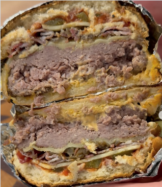 Nahaufnahme eines halbierten Burgers, bei dem beide Hälften übereinander liegen. Sichtbar sind mehrere Schichten: ein dickes Rindfleisch-Patty, geschmolzener Käse, Gurkenscheiben, Zwiebeln und Soße, umhüllt von einem knusprigen Brötchen. Der Burger ist in Folie eingewickelt.