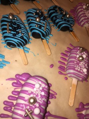 Mehrere dekorierte Cake-Pops auf Holzstielen, teilweise mit blauer und schwarzer Glasur sowie silbernen Zuckerperlen verziert, daneben Varianten mit weißer und pinker Glasur, garniert mit weißen Streuseln und silbernen Dekorelementen, auf braunem Backpapier.