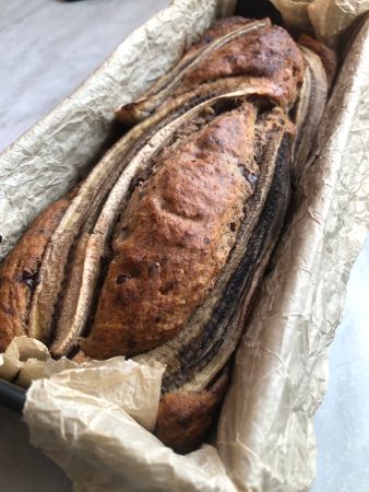Frisch gebackenes Bananenbrot in einer mit Backpapier ausgelegten Kastenform. Das Brot ist goldbraun und oben mit längs halbierten Bananen dekoriert.