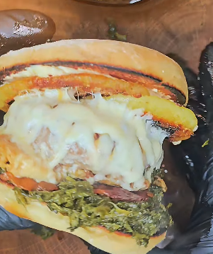 Nahaufnahme eines Burgers mit geschmolzenem Käse, Grünkohl, Fleisch und weiteren Zutaten zwischen zwei gerösteten Brötchenhälften. Der Burger wird von einer Hand mit schwarzem Handschuh gehalten.