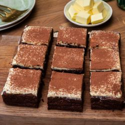 Auf einem Holzbrett liegen neun Brownies mit heller Creme und einer Schicht Kakaopulver. Daneben stehen ein feines Sieb, ein kleiner Teller mit Kakaopulver und ein Teller mit Butterwürfeln.