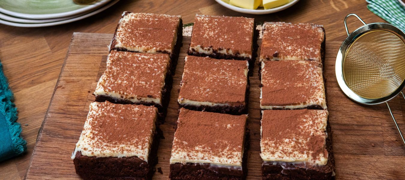 Auf einem Holzbrett liegen neun Brownies mit heller Creme und einer Schicht Kakaopulver. Daneben stehen ein feines Sieb, ein kleiner Teller mit Kakaopulver und ein Teller mit Butterwürfeln.