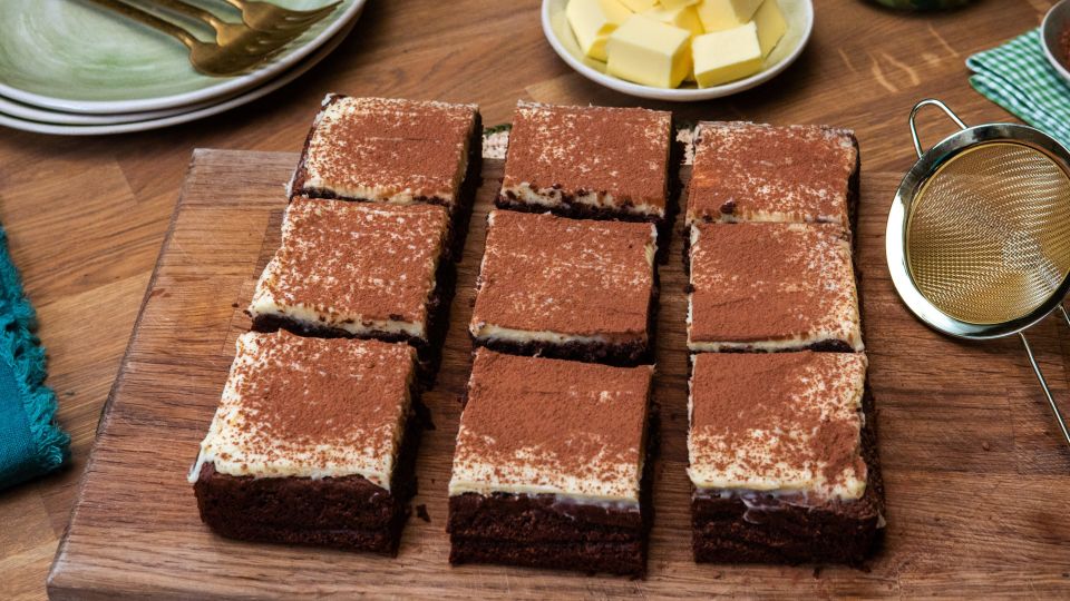 Auf einem Holzbrett liegen neun Brownies mit heller Creme und einer Schicht Kakaopulver. Daneben stehen ein feines Sieb, ein kleiner Teller mit Kakaopulver und ein Teller mit Butterwürfeln.