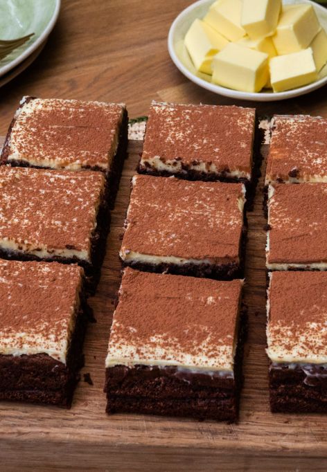 Auf einem Holzbrett liegen neun Brownies mit heller Creme und einer Schicht Kakaopulver. Daneben stehen ein feines Sieb, ein kleiner Teller mit Kakaopulver und ein Teller mit Butterwürfeln.