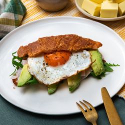 Auf einem weißen Teller liegt ein halbiertes Croissant, gefüllt mit Avocado, Rucola, Schinken und einem Spiegelei.