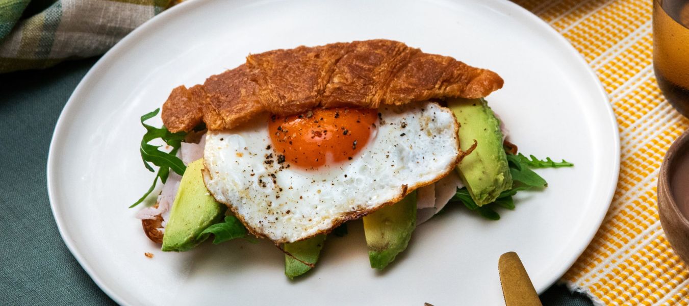 Auf einem weißen Teller liegt ein halbiertes Croissant, gefüllt mit Avocado, Rucola, Schinken und einem Spiegelei.