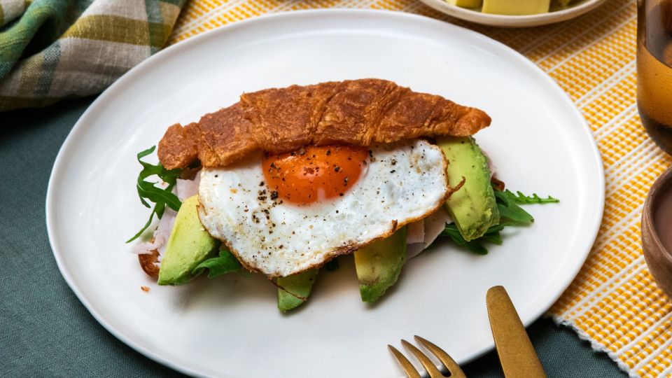 Auf einem weißen Teller liegt ein halbiertes Croissant, gefüllt mit Avocado, Rucola, Schinken und einem Spiegelei.