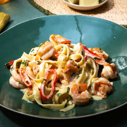 Ein grüner Teller mit Tagliatelle in cremiger Sauce, serviert mit Garnelen, roten Chilistreifen und frischer Petersilie.