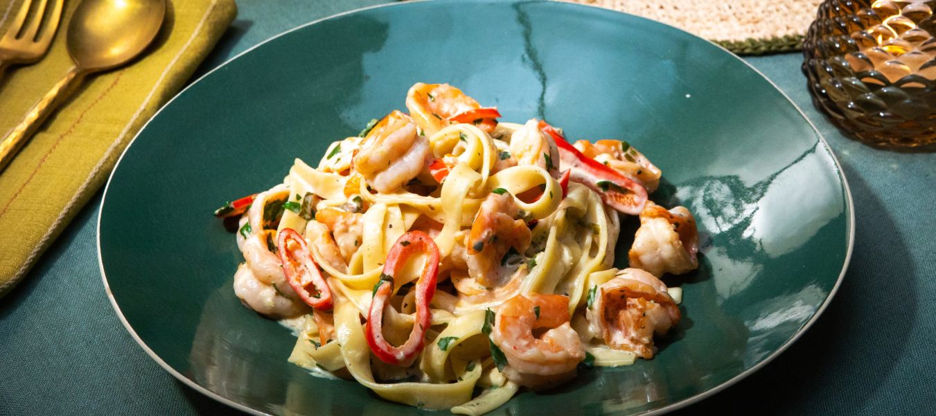 Ein grüner Teller mit Tagliatelle in cremiger Sauce, serviert mit Garnelen, roten Chilistreifen und frischer Petersilie.