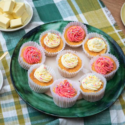 Ein großer grüner Teller mit mehreren Cupcakes, verziert mit rosafarbenen und heller Buttercreme sowie bunten Zuckerstreuseln.