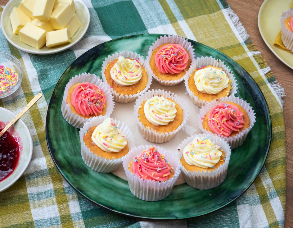Ein großer grüner Teller mit mehreren Cupcakes, verziert mit rosafarbenen und heller Buttercreme sowie bunten Zuckerstreuseln.