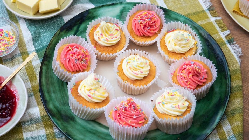 Ein großer grüner Teller mit mehreren Cupcakes, verziert mit rosafarbenen und heller Buttercreme sowie bunten Zuckerstreuseln.