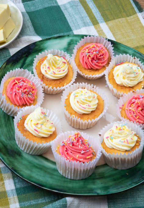 Ein großer grüner Teller mit mehreren Cupcakes, verziert mit rosafarbenen und heller Buttercreme sowie bunten Zuckerstreuseln.