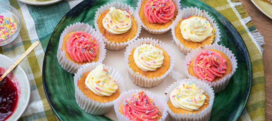 Ein großer grüner Teller mit mehreren Cupcakes, verziert mit rosafarbenen und heller Buttercreme sowie bunten Zuckerstreuseln.