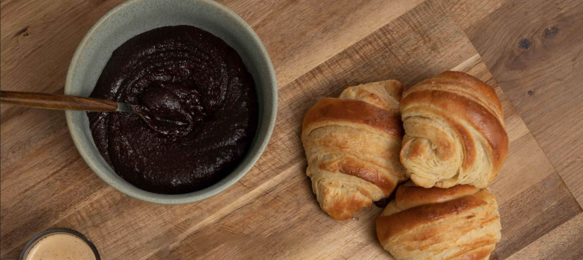 Eine Schale mit dunkler, cremiger Schokoladencreme und einem Holzlöffel. Daneben liegen drei goldbraun gebackene Croissants auf einer Holzoberfläche.