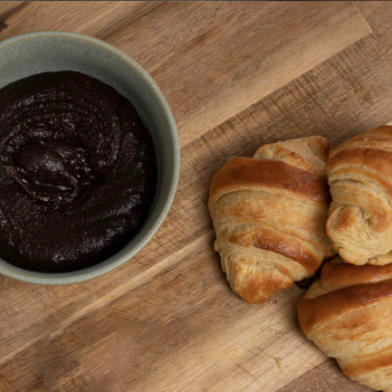 Eine Schale mit dunkler, cremiger Schokoladencreme und einem Holzlöffel. Daneben liegen drei goldbraun gebackene Croissants auf einer Holzoberfläche.
