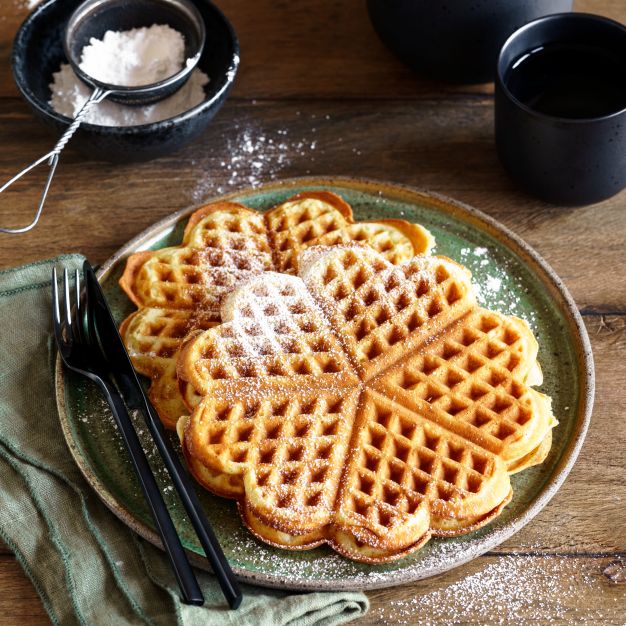 Ein Teller mit herzförmigen Waffeln, bestäubt mit Puderzucker, steht auf einem rustikalen Holztisch. Daneben stehen eine schwarze Teekanne, eine schwarze Tasse, Gabeln und eine grüne Serviette. Daneben steht ein Sieb mit Puderzucker.