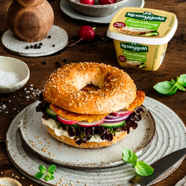 Ein Sesam-Hähnchen-Bagel-Sandwich gefüllt mit Salat, Tomaten, Gurken, roten Zwiebeln und einem goldbraun panierten Hähnchen-Patty liegt auf einem Keramikteller, umgeben von frischen Radieschen, Basilikum, Gewürzen und einer Salzmühle auf einem Holztisch.