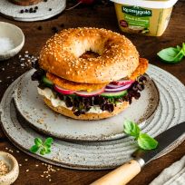 Ein Sesam-Hähnchen-Bagel-Sandwich gefüllt mit Salat, Tomaten, Gurken, roten Zwiebeln und einem goldbraun panierten Hähnchen-Patty liegt auf einem Keramikteller, umgeben von frischen Radieschen, Basilikum, Gewürzen und einer Salzmühle auf einem Holztisch.
