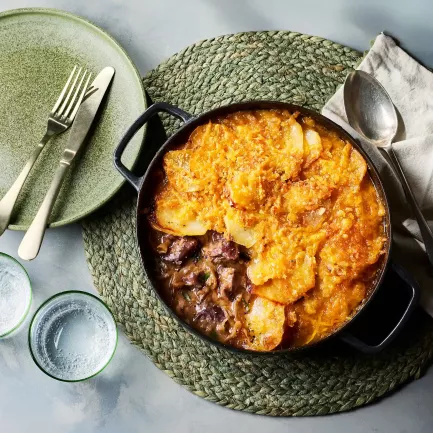 Ein gusseiserner Topf mit einem Lammeintopf steht auf einem runden, geflochtenen Untersetzer. Obenauf liegt eine goldbraun gebackene Schicht aus Kartoffeln und Käse. Daneben stehen ein grüner Teller mit Besteck, zwei Trinkgläser sowie eine Serviette.