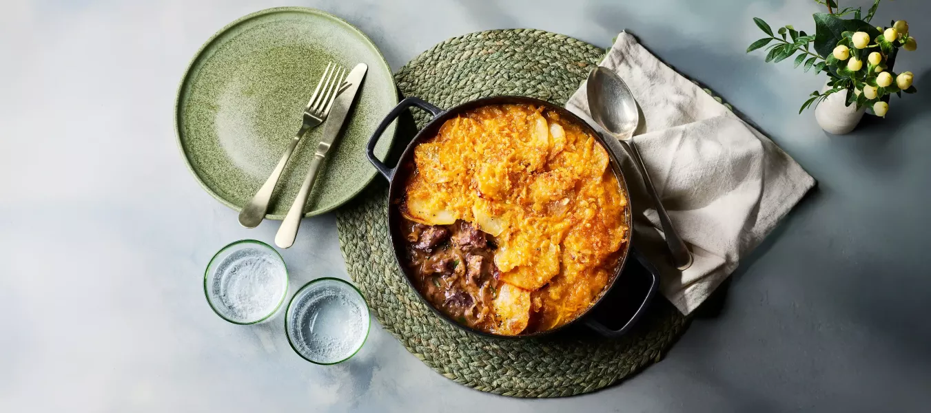 Ein gusseiserner Topf mit einem Lammeintopf steht auf einem runden, geflochtenen Untersetzer. Obenauf liegt eine goldbraun gebackene Schicht aus Kartoffeln und Käse. Daneben stehen ein grüner Teller mit Besteck, zwei Trinkgläser sowie eine Serviette.
