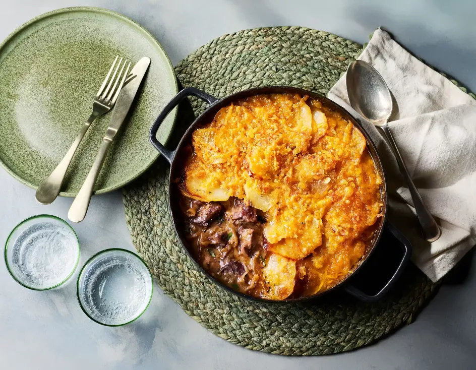 Ein gusseiserner Topf mit einem Lammeintopf steht auf einem runden, geflochtenen Untersetzer. Obenauf liegt eine goldbraun gebackene Schicht aus Kartoffeln und Käse. Daneben stehen ein grüner Teller mit Besteck, zwei Trinkgläser sowie eine Serviette.