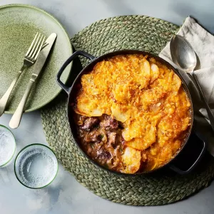 Ein gusseiserner Topf mit einem Lammeintopf steht auf einem runden, geflochtenen Untersetzer. Obenauf liegt eine goldbraun gebackene Schicht aus Kartoffeln und Käse. Daneben stehen ein grüner Teller mit Besteck, zwei Trinkgläser sowie eine Serviette.