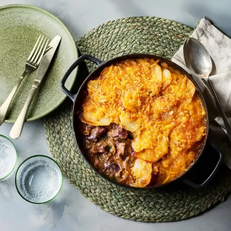 Ein gusseiserner Topf mit einem Lammeintopf steht auf einem runden, geflochtenen Untersetzer. Obenauf liegt eine goldbraun gebackene Schicht aus Kartoffeln und Käse. Daneben stehen ein grüner Teller mit Besteck, zwei Trinkgläser sowie eine Serviette.