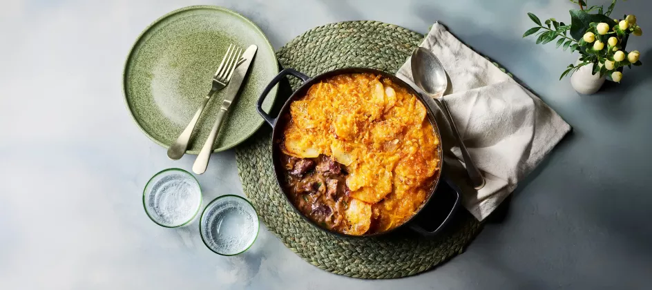 Ein gusseiserner Topf mit einem Lammeintopf steht auf einem runden, geflochtenen Untersetzer. Obenauf liegt eine goldbraun gebackene Schicht aus Kartoffeln und Käse. Daneben stehen ein grüner Teller mit Besteck, zwei Trinkgläser sowie eine Serviette.