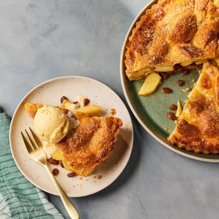Eine goldbraun gebackene Apfel-Whiskey‑Tarte mit knuspriger Zuckerkruste steht auf einem grünen Teller. Ein Stück wurde herausgeschnitten, sodass die Apfel‑Whiskey‑Füllung sichtbar wird. Daneben liegt ein Teller mit einem Stück der Tarte, serviert mit einer Kugel Eis und einigen Rosinen.