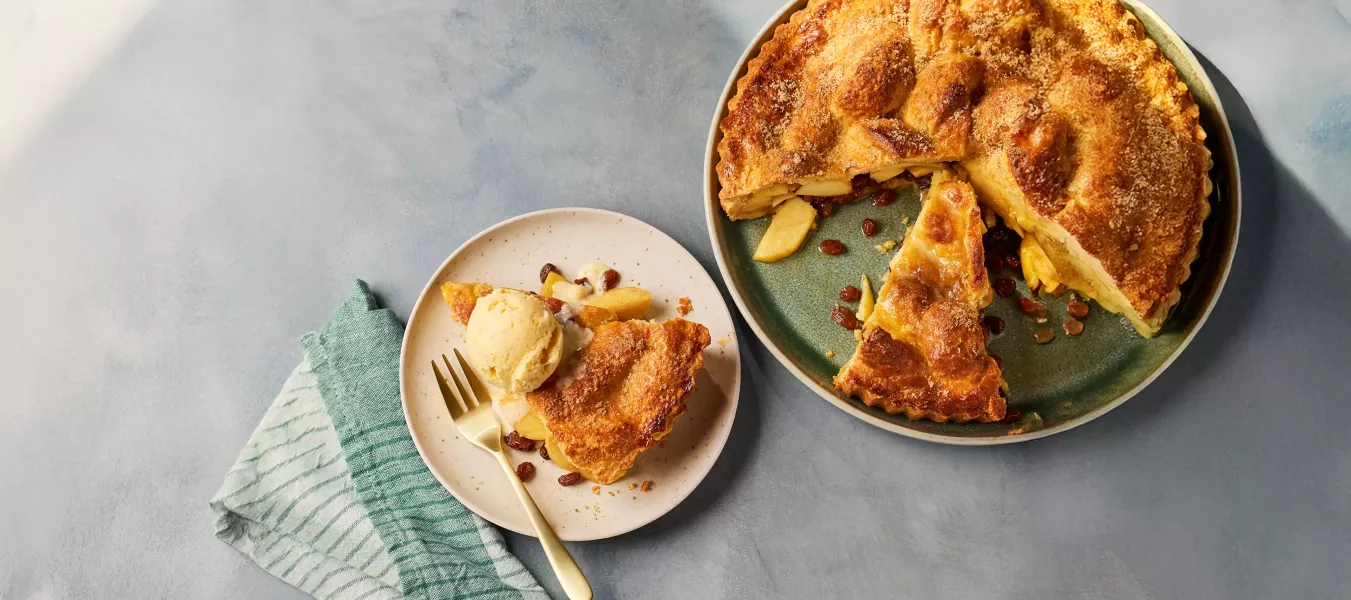 Eine goldbraun gebackene Apfel-Whiskey‑Tarte mit knuspriger Zuckerkruste steht auf einem grünen Teller. Ein Stück wurde herausgeschnitten, sodass die Apfel‑Whiskey‑Füllung sichtbar wird. Daneben liegt ein Teller mit einem Stück der Tarte, serviert mit einer Kugel Eis und einigen Rosinen.