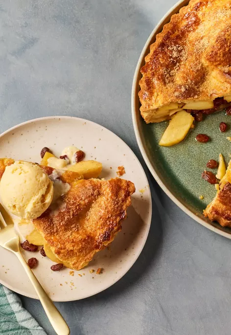 Eine goldbraun gebackene Apfel-Whiskey‑Tarte mit knuspriger Zuckerkruste steht auf einem grünen Teller. Ein Stück wurde herausgeschnitten, sodass die Apfel‑Whiskey‑Füllung sichtbar wird. Daneben liegt ein Teller mit einem Stück der Tarte, serviert mit einer Kugel Eis und einigen Rosinen.