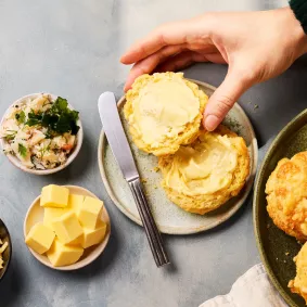 Mehrere goldbraun gebackene, käsige Scones liegen auf einem Teller. Ein aufgeschnittener Scone wird mit Butter bestrichen, daneben stehen kleine Schalen mit Käsewürfeln, geriebenem Käse und einer Krabbenmischung.