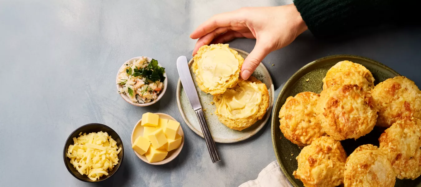 Mehrere goldbraun gebackene, käsige Scones liegen auf einem Teller. Ein aufgeschnittener Scone wird mit Butter bestrichen, daneben stehen kleine Schalen mit Käsewürfeln, geriebenem Käse und einer Krabbenmischung.