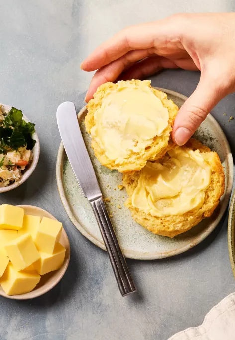 Mehrere goldbraun gebackene, käsige Scones liegen auf einem Teller. Ein aufgeschnittener Scone wird mit Butter bestrichen, daneben stehen kleine Schalen mit Käsewürfeln, geriebenem Käse und einer Krabbenmischung.