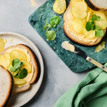 Zwei belegte Sandwiches liegen auf einem hellen Teller und einem grünen Schneidebrett. Die Brotscheiben sind mit Butter bestrichen, darauf liegen Chips und etwas Kresse.