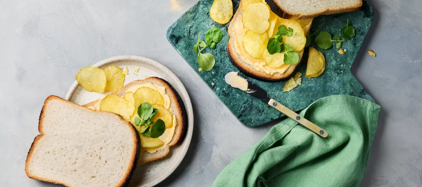 Zwei belegte Sandwiches liegen auf einem hellen Teller und einem grünen Schneidebrett. Die Brotscheiben sind mit Butter bestrichen, darauf liegen Chips und etwas Kresse.