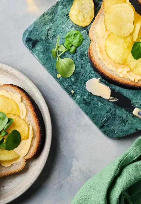 Zwei belegte Sandwiches liegen auf einem hellen Teller und einem grünen Schneidebrett. Die Brotscheiben sind mit Butter bestrichen, darauf liegen Chips und etwas Kresse.