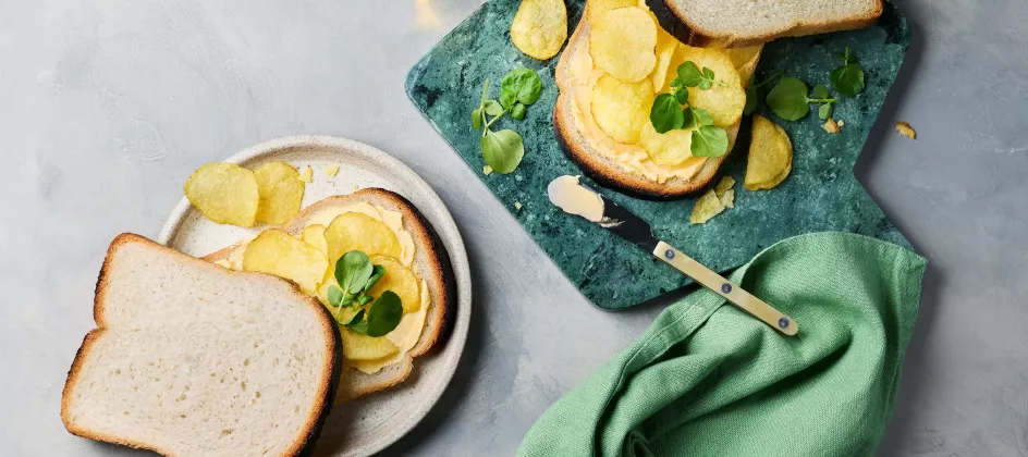 Zwei belegte Sandwiches liegen auf einem hellen Teller und einem grünen Schneidebrett. Die Brotscheiben sind mit Butter bestrichen, darauf liegen Chips und etwas Kresse.