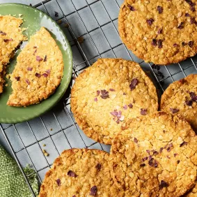 Mehrere runde, goldbraun gebackene Hafercookies liegen auf einem Abkühlgitter. Ein Teller mit einem in zwei Hälften gebrochenen Cookie steht daneben.
